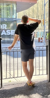 A person stands on a terrace, gazing out through a metal railing at a city street. They are wearing a black t-shirt, dark shorts, and colorful shoes. A large building with glass windows and an 'For Lease' sign is visible in the background, along with orange traffic cones.