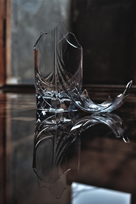 A broken glass object resting on a reflective surface with shards scattered around. The surroundings are dimly lit, giving the image a somber and contemplative feel.