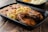 A black plastic food container holds a serving of rice and beans, macaroni salad with vegetables, and two pieces of well-seasoned fried chicken. The food is arranged neatly against a rustic wooden table background.