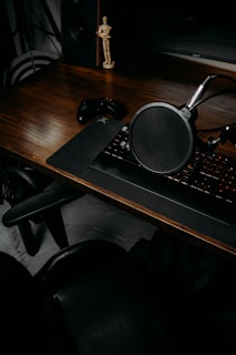A wooden desk is arranged with a microphone on a flexible arm, a keyboard on a large mouse pad, a gaming controller, and a small mannequin figure placed next to a speaker. The setup suggests a workspace or gaming area, with dark furniture and muted lighting creating a cozy atmosphere.