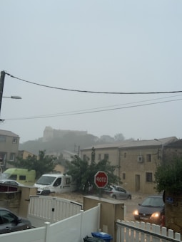 A residential street appears under heavy rain or a storm. Several vehicles, including a van, are parked or driving on a road partially covered in water. A stop sign is visible, and the surrounding houses have a rustic appearance with a hill and large structure in the background, partially obscured by fog or rain.
