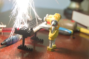 A small humanoid figure made from mechanical parts appears to be welding or working with a metal piece on a table. Bright sparks fly upward from the contact point, illuminating the scene. The background includes various tools and a book, set on a wooden surface.