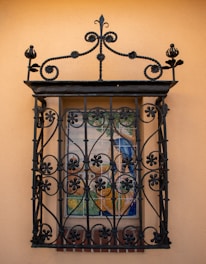 An ornate black wrought iron grille covers a small window, which features a colorful detailed mural. The mural depicts a serene outdoor scene with trees and possibly a building in the background. The wall is a warm beige color, complementing the dark ironwork.