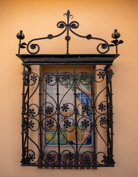 An ornate black wrought iron grille covers a small window, which features a colorful detailed mural. The mural depicts a serene outdoor scene with trees and possibly a building in the background. The wall is a warm beige color, complementing the dark ironwork.