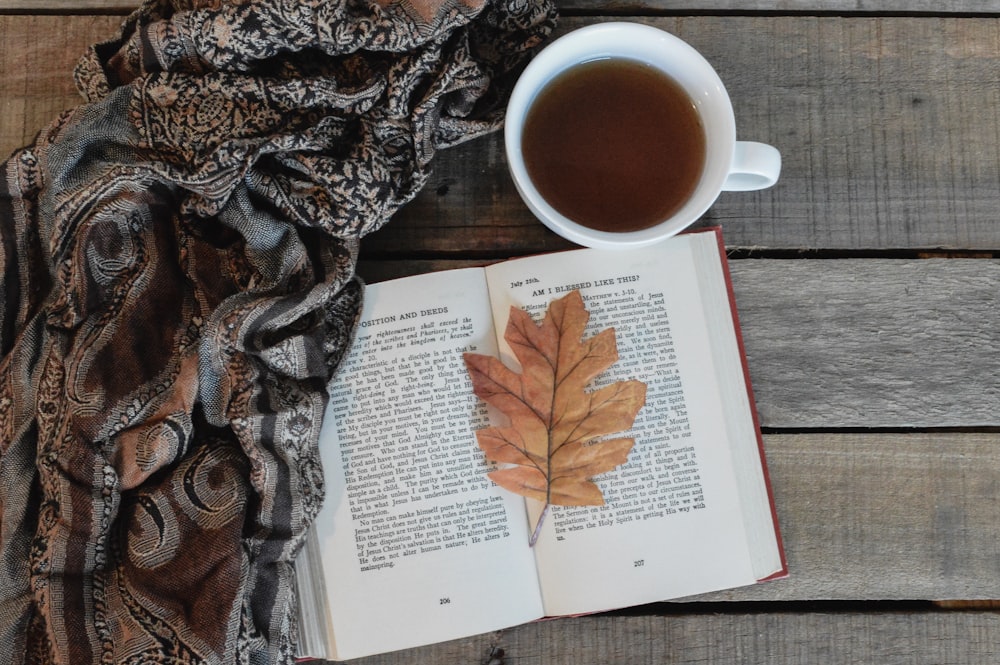 an open book with a leaf on top of it next to a cup of coffee