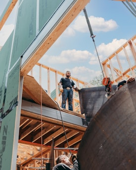 Construction workers are engaged in building a wooden framework structure. One worker stands on the upper level, while another is positioned on a lower level, installing materials. A crane is visible, lifting a large object. The sky is clear with some clouds.