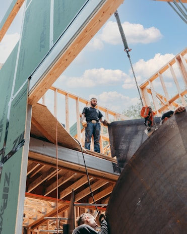 Construction workers are engaged in building a wooden framework structure. One worker stands on the upper level, while another is positioned on a lower level, installing materials. A crane is visible, lifting a large object. The sky is clear with some clouds.