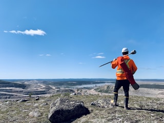 A person in high-visibility orange safety gear and a white helmet stands on a rocky hill, overlooking a vast landscape with a quarry or mining operation and a river. The blue sky is clear, with a few scattered clouds.
