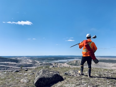 A person in high-visibility orange safety gear and a white helmet stands on a rocky hill, overlooking a vast landscape with a quarry or mining operation and a river. The blue sky is clear, with a few scattered clouds.