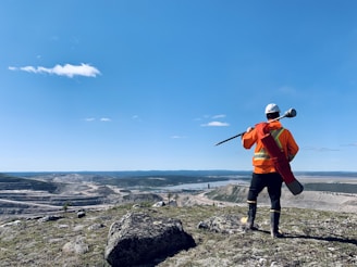 A person in high-visibility orange safety gear and a white helmet stands on a rocky hill, overlooking a vast landscape with a quarry or mining operation and a river. The blue sky is clear, with a few scattered clouds.