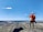 A person in high-visibility orange safety gear and a white helmet stands on a rocky hill, overlooking a vast landscape with a quarry or mining operation and a river. The blue sky is clear, with a few scattered clouds.