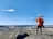 A person in high-visibility orange safety gear and a white helmet stands on a rocky hill, overlooking a vast landscape with a quarry or mining operation and a river. The blue sky is clear, with a few scattered clouds.