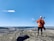 A person in high-visibility orange safety gear and a white helmet stands on a rocky hill, overlooking a vast landscape with a quarry or mining operation and a river. The blue sky is clear, with a few scattered clouds.