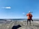 A person in high-visibility orange safety gear and a white helmet stands on a rocky hill, overlooking a vast landscape with a quarry or mining operation and a river. The blue sky is clear, with a few scattered clouds.