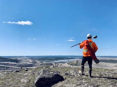 A person in high-visibility orange safety gear and a white helmet stands on a rocky hill, overlooking a vast landscape with a quarry or mining operation and a river. The blue sky is clear, with a few scattered clouds.