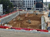 A construction site with heavy machinery working on a large excavation. There are several excavators and bulldozers actively moving earth within a fenced-off area. Surrounding the site are buildings under construction and various construction materials. The scene is bustling with activity, indicating progress in the development project.