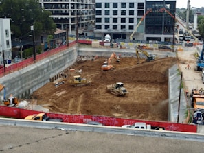 A construction site with heavy machinery working on a large excavation. There are several excavators and bulldozers actively moving earth within a fenced-off area. Surrounding the site are buildings under construction and various construction materials. The scene is bustling with activity, indicating progress in the development project.