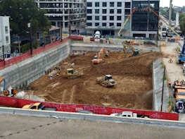 A construction site with heavy machinery working on a large excavation. There are several excavators and bulldozers actively moving earth within a fenced-off area. Surrounding the site are buildings under construction and various construction materials. The scene is bustling with activity, indicating progress in the development project.