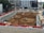 A construction site with heavy machinery working on a large excavation. There are several excavators and bulldozers actively moving earth within a fenced-off area. Surrounding the site are buildings under construction and various construction materials. The scene is bustling with activity, indicating progress in the development project.