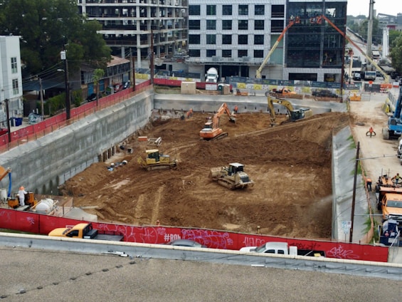 A construction site with heavy machinery working on a large excavation. There are several excavators and bulldozers actively moving earth within a fenced-off area. Surrounding the site are buildings under construction and various construction materials. The scene is bustling with activity, indicating progress in the development project.