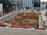 A construction site with heavy machinery working on a large excavation. There are several excavators and bulldozers actively moving earth within a fenced-off area. Surrounding the site are buildings under construction and various construction materials. The scene is bustling with activity, indicating progress in the development project.