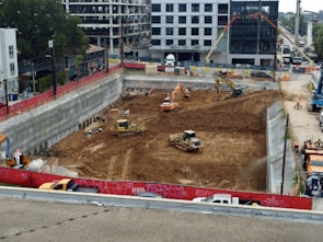 A construction site with heavy machinery working on a large excavation. There are several excavators and bulldozers actively moving earth within a fenced-off area. Surrounding the site are buildings under construction and various construction materials. The scene is bustling with activity, indicating progress in the development project.
