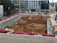 A construction site with heavy machinery working on a large excavation. There are several excavators and bulldozers actively moving earth within a fenced-off area. Surrounding the site are buildings under construction and various construction materials. The scene is bustling with activity, indicating progress in the development project.