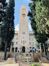 A tall, beige stone building with an ornate architectural design, featuring a prominent central tower. The entrance is flanked by two tall trees, and the area is landscaped with steps leading to the building. There are signs that identify the location as a YMCA.