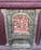 An intricately carved relief featuring human figures in traditional attire, surrounded by ornate designs. The central figure appears to be raised on a platform or altar, with others beside and around them in various poses. The relief is framed by a decorative border with floral and scroll patterns.