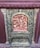 An intricately carved relief featuring human figures in traditional attire, surrounded by ornate designs. The central figure appears to be raised on a platform or altar, with others beside and around them in various poses. The relief is framed by a decorative border with floral and scroll patterns.