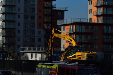 A yellow excavator is positioned in front of modern residential buildings with multiple balconies. The structures are high-rise apartments, featuring a mix of gray and orange exteriors. The setting appears urban, with the excavator likely involved in construction or maintenance work.