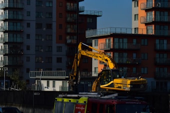 A yellow excavator is positioned in front of modern residential buildings with multiple balconies. The structures are high-rise apartments, featuring a mix of gray and orange exteriors. The setting appears urban, with the excavator likely involved in construction or maintenance work.