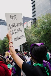 A person in a crowd holds up a sign with text written in Spanish, surrounded by tall buildings and trees. The atmosphere appears to be that of a protest or rally, with people wearing scarves or bandanas, some in purple and green.