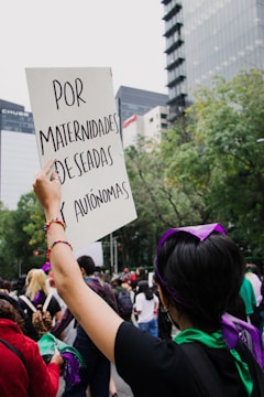A person in a crowd holds up a sign with text written in Spanish, surrounded by tall buildings and trees. The atmosphere appears to be that of a protest or rally, with people wearing scarves or bandanas, some in purple and green.