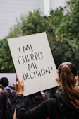 A person holds up a sign with the text '¡MI CUERPO, MI DECISIÓN!' in a crowd. The background shows trees and a building, indicating an outdoor setting. The focus is on the sign, which is prominently displayed above the person's head.