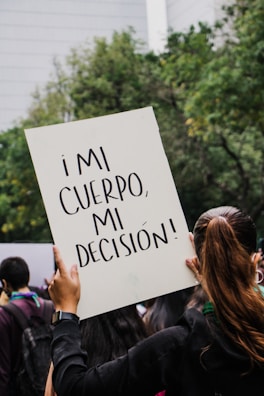 A person holds up a sign with the text '¡MI CUERPO, MI DECISIÓN!' in a crowd. The background shows trees and a building, indicating an outdoor setting. The focus is on the sign, which is prominently displayed above the person's head.