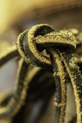 Close-up of a textured, frayed black and yellow shoelace, intricately tied in a knot. The image captures the rugged surface with visible dust particles, creating a tactile and worn appearance. The background is softly blurred, drawing focus to the shoelace.