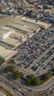 An aerial view of a large commercial shopping complex surrounded by a densely packed parking lot filled with cars. The complex has multiple large buildings with beige rooftops, and nearby streets are lined with smaller residential buildings. Trees and landscaped areas are visible around the edges, alongside busy roads and vehicles.