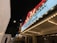 A night scene of a theater marquee illuminated by bright lights. The marquee displays text about a comedy festival and a performance at a venue called 'Your Mom’s House.' Additional lights and signage are visible in the background, contributing to an urban, lively atmosphere.