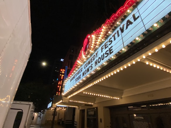 A night scene of a theater marquee illuminated by bright lights. The marquee displays text about a comedy festival and a performance at a venue called 'Your Mom’s House.' Additional lights and signage are visible in the background, contributing to an urban, lively atmosphere.