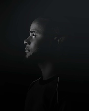 A profile view of a person with a contemplative expression set against a dark background. The lighting highlights the facial features while the rest of the image remains in shadows, emphasizing the subject's thoughtful demeanor.