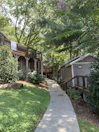 A pathway lined with greenery leads to wooden buildings surrounded by lush trees. Towels hang over the railing of an elevated wooden deck that is part of one of the buildings. The scene is peaceful and shaded by tall trees, with some small flowering plants visible.