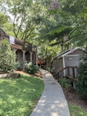 A pathway lined with greenery leads to wooden buildings surrounded by lush trees. Towels hang over the railing of an elevated wooden deck that is part of one of the buildings. The scene is peaceful and shaded by tall trees, with some small flowering plants visible.