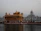 The image depicts a stunning architectural marvel with a golden temple structure on a serene body of water. The temple showcases intricate details and is surrounded by a harmonious blend of traditional and modern architectural elements. The backdrop features a white dome-laden building, adding to the spiritual and tranquil ambiance.