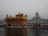 The image depicts a stunning architectural marvel with a golden temple structure on a serene body of water. The temple showcases intricate details and is surrounded by a harmonious blend of traditional and modern architectural elements. The backdrop features a white dome-laden building, adding to the spiritual and tranquil ambiance.