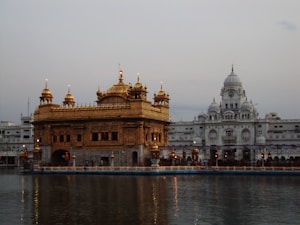 The image depicts a stunning architectural marvel with a golden temple structure on a serene body of water. The temple showcases intricate details and is surrounded by a harmonious blend of traditional and modern architectural elements. The backdrop features a white dome-laden building, adding to the spiritual and tranquil ambiance.