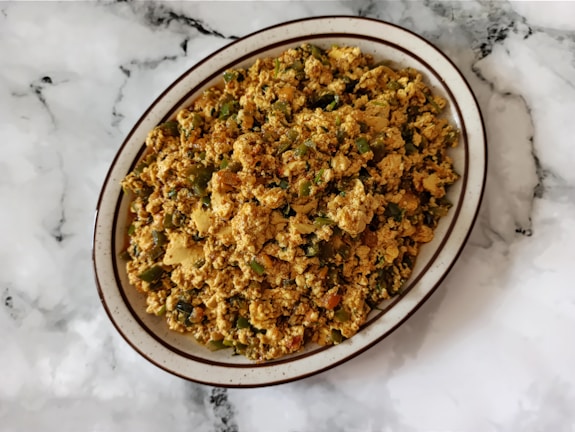 A plate of scrambled tofu with vegetables is placed on a marbled surface. The dish contains a mix of crumbled tofu, green bell peppers, onions, and possibly spices, creating a colorful and textured appearance.