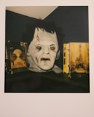 A mask or bust resembling a classic horror movie character is positioned between two vintage horror movie covers or posters. The face has a pale complexion, with sunken eyes and a noticeable scar on the forehead. The surrounding lighting is dim, enhancing the eerie appearance of the mask. The items are placed on a dark surface under a slanted ceiling.