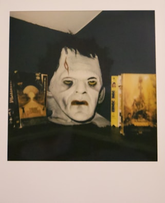 A mask or bust resembling a classic horror movie character is positioned between two vintage horror movie covers or posters. The face has a pale complexion, with sunken eyes and a noticeable scar on the forehead. The surrounding lighting is dim, enhancing the eerie appearance of the mask. The items are placed on a dark surface under a slanted ceiling.