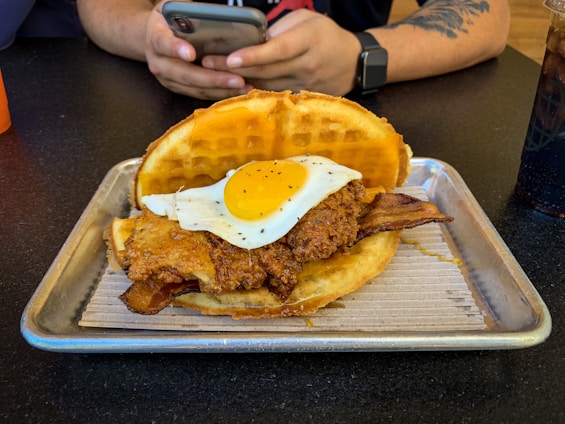 A waffle sandwich on a metal tray contains fried chicken topped with a sunny-side-up egg and bacon. A person with tattooed arms is seated at a table in the background, holding a smartphone. A drink is positioned to the right.
